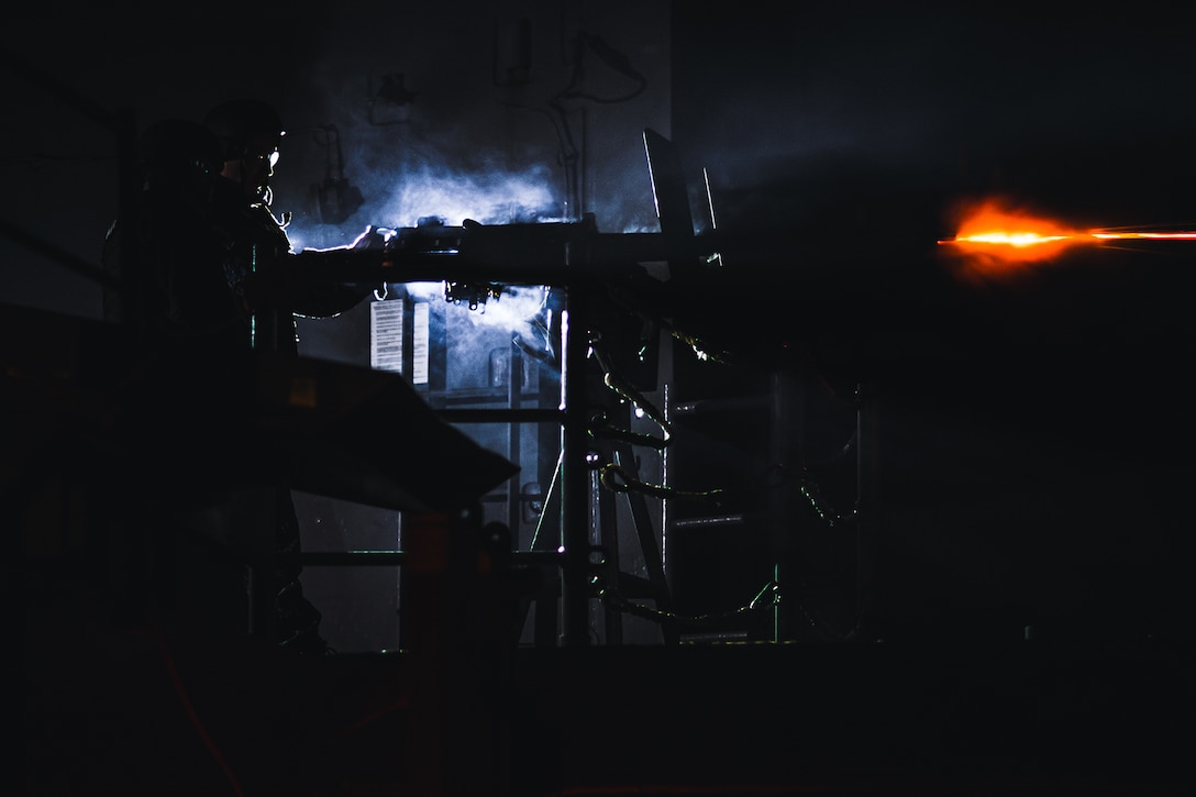 An orange flame is seen in the dark as a sailor in silhouette fires a machine gun aboard a ship at night.