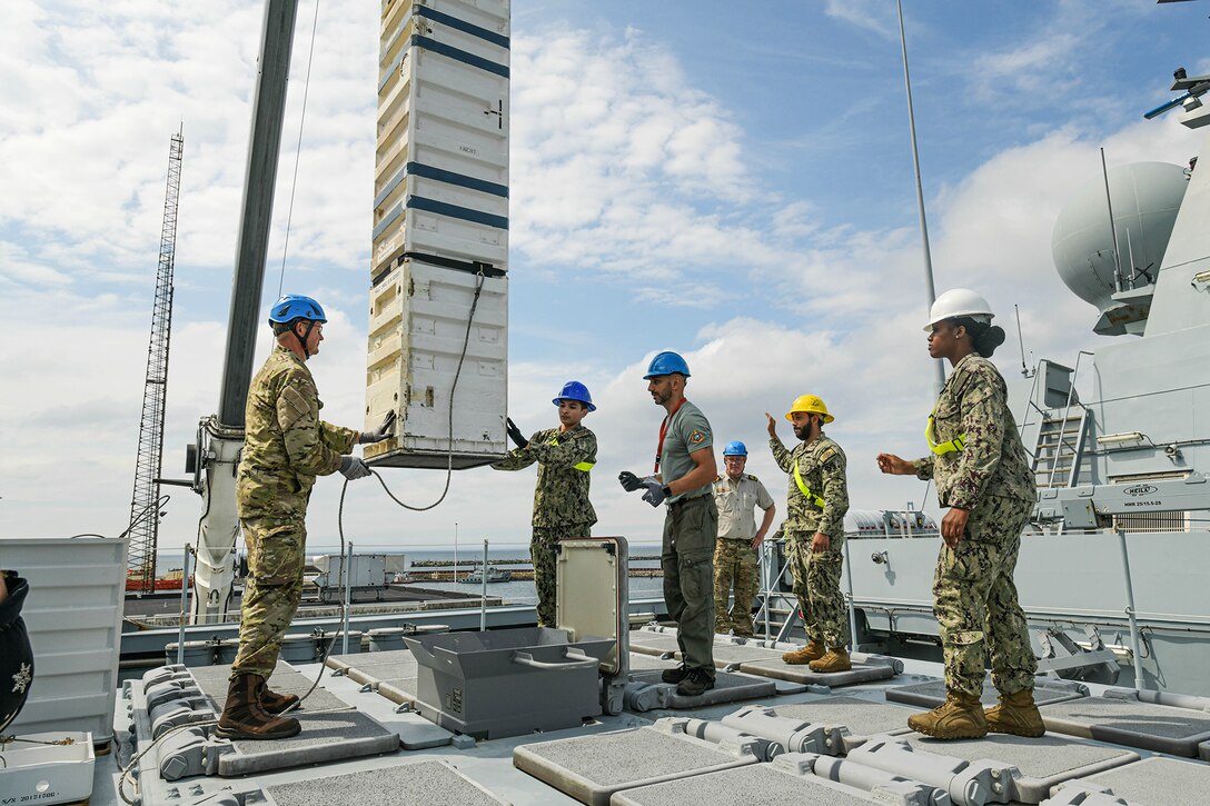 A half-dozen U.S. and Danish sailors stand on a ship's deck, two of them handling a tall object hoisted in the air.