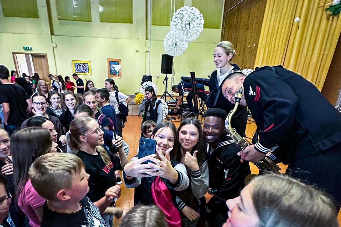 Navy musicians pose for a selfie with young people standing among other audience members.