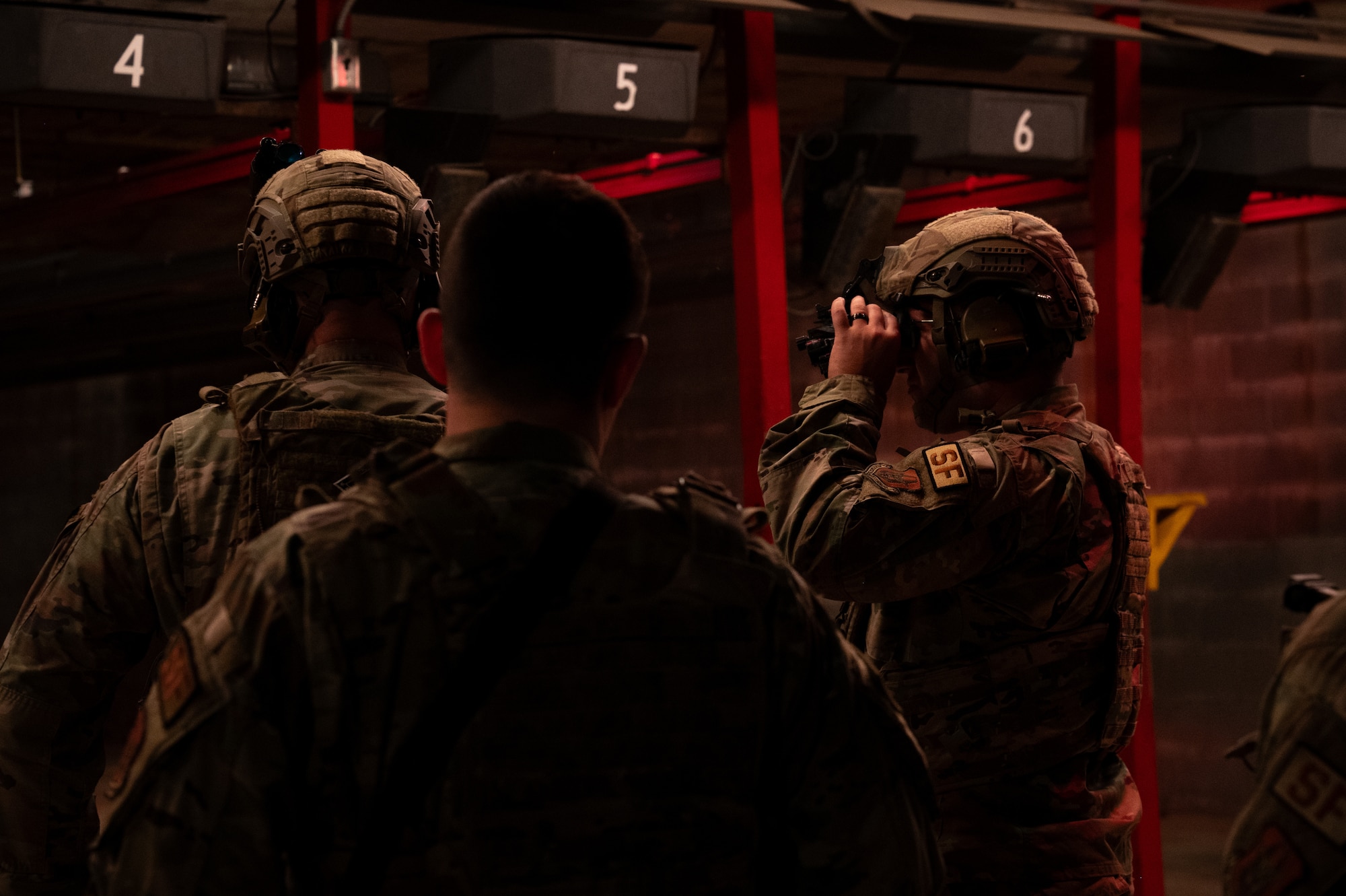 Backs of heads and side profiles of uniformed military members. Dark lighting encompasses the photo with red lights in the background of the image, behind the service members.