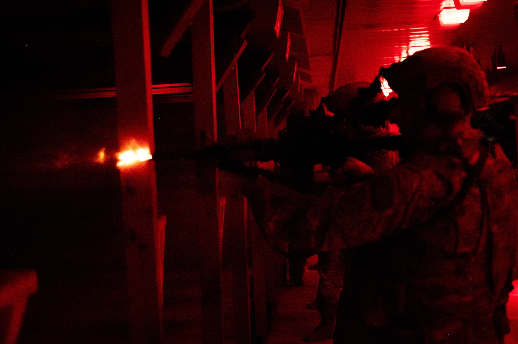 A red and black image that shows the side profile of a military member holding a weapon. At the tip of the weapon, a flash of light that indicates a bullet has been fired.