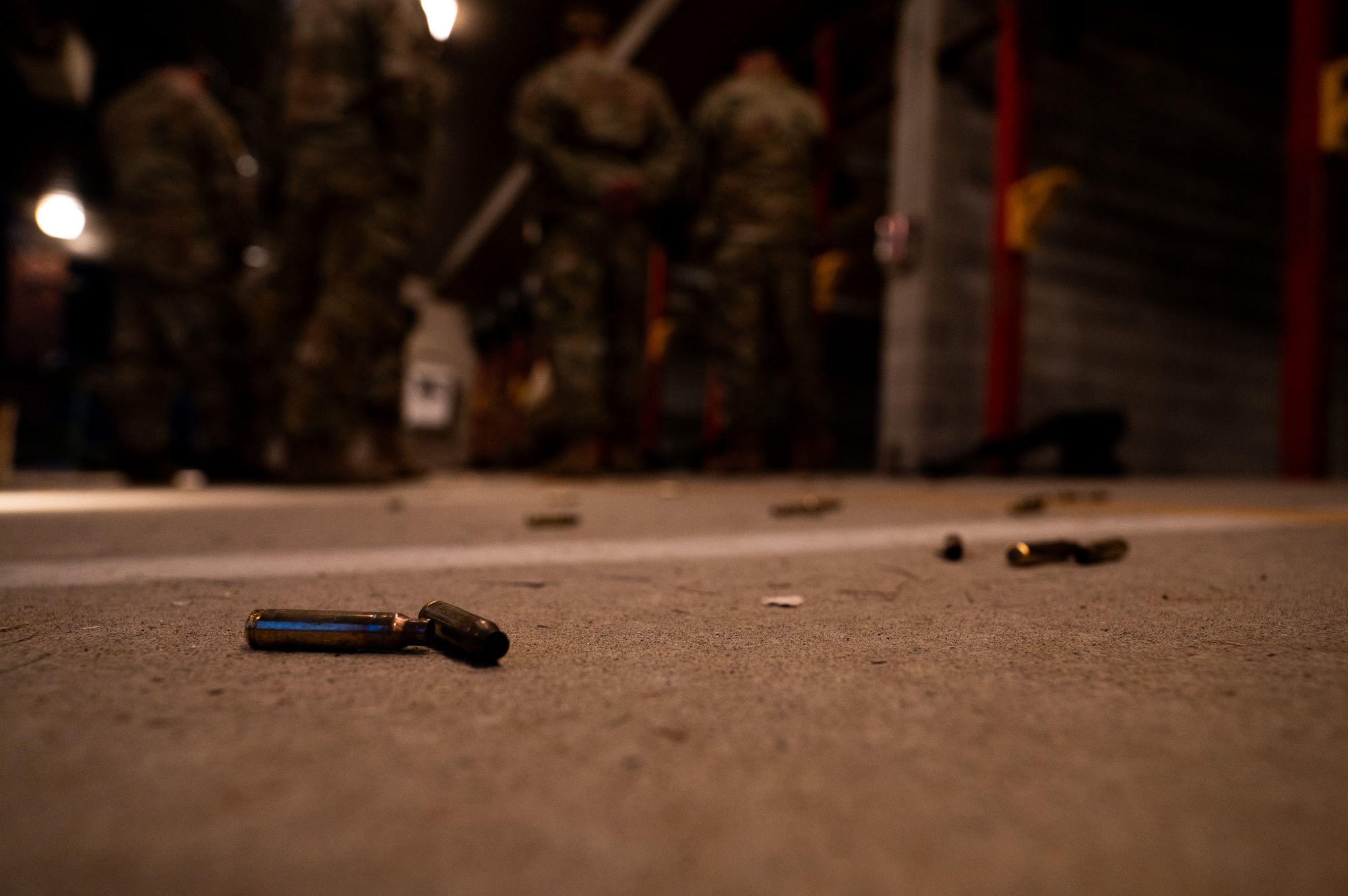 Stray bullet casings litter the ground. In the background, a white line painted on the concrete and a gun range visible.