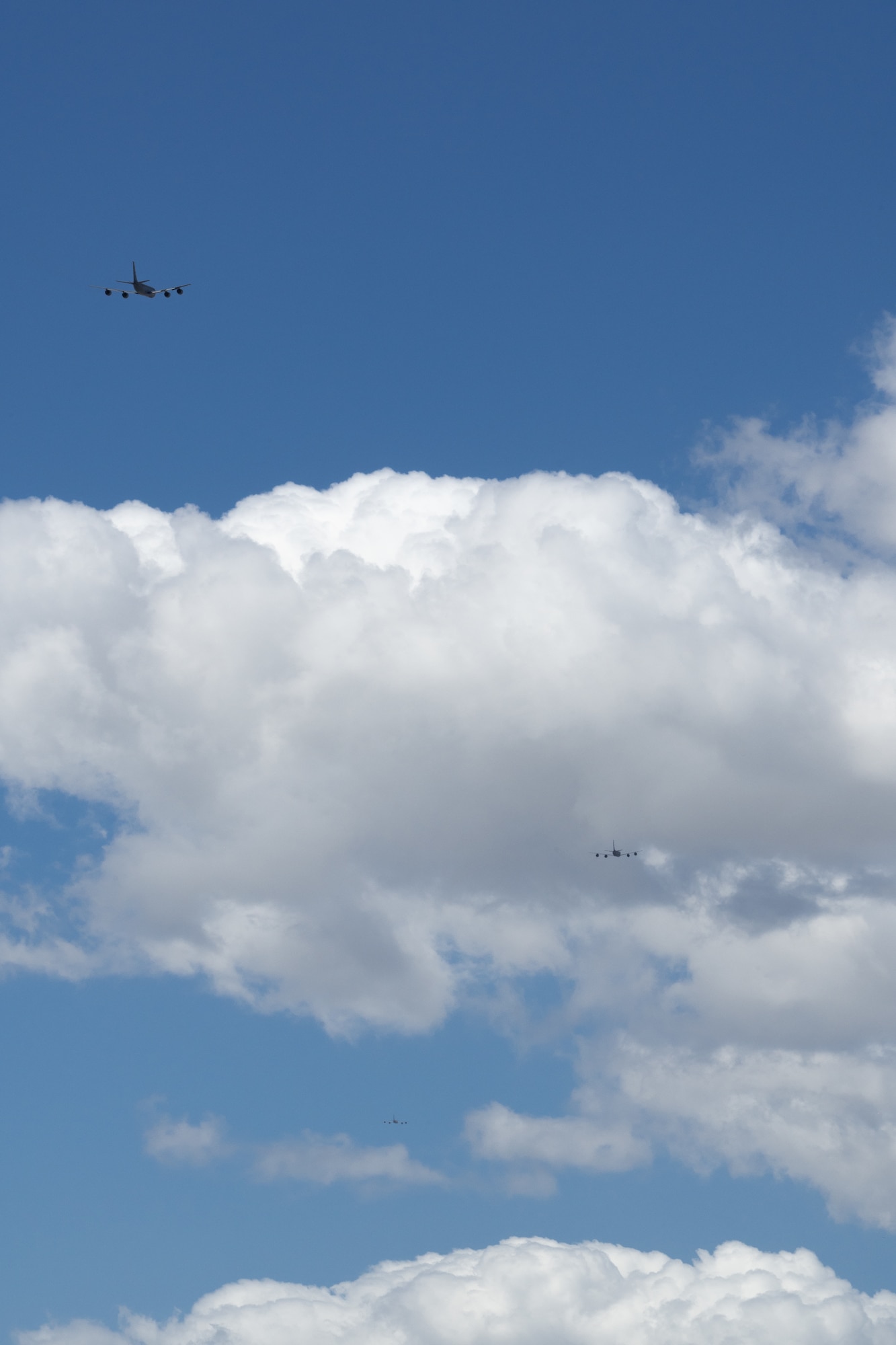 Blue sky and white clouds fill the frame. In the top left corner, the tail of an aircraft. In the middle of a cloud at right, another. In the far distance, lowest in the photograph, a glimpse of an aircraft is visible. All three are gray in color.