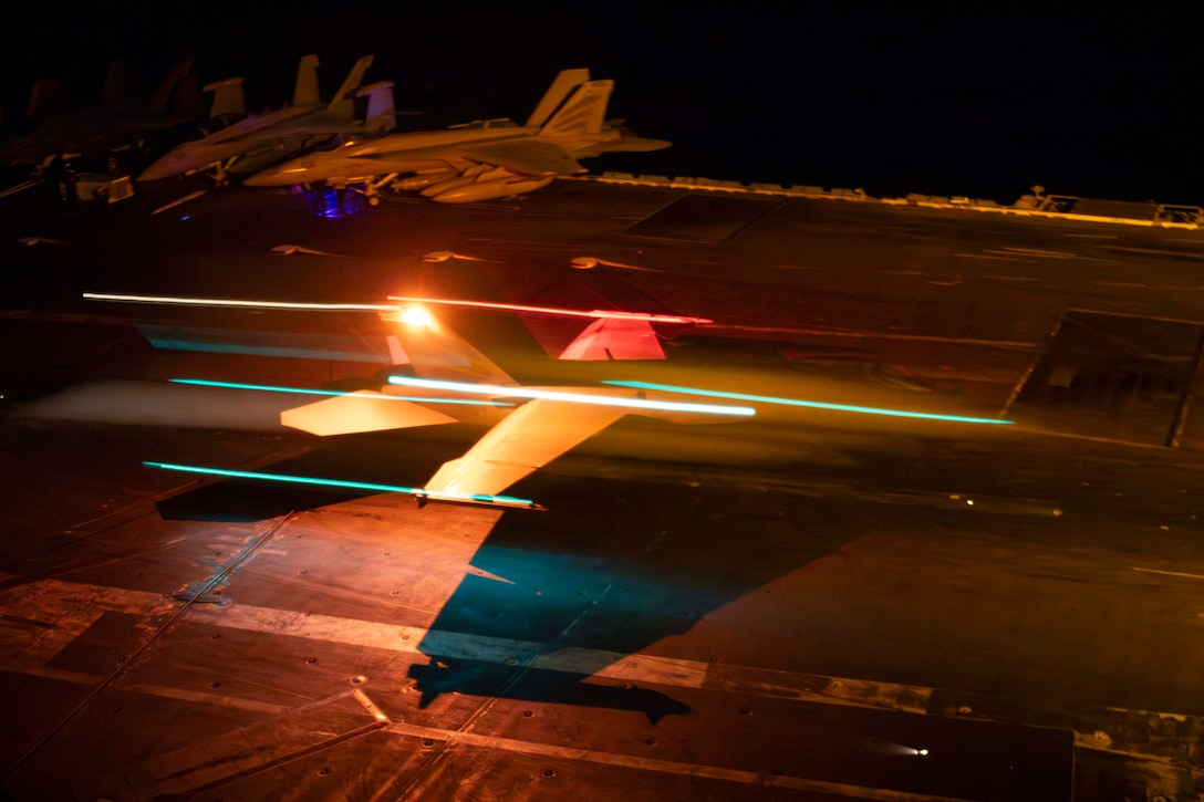 Overhead view of a military aircraft in motion, landing on the deck of a Navy ship at night.
