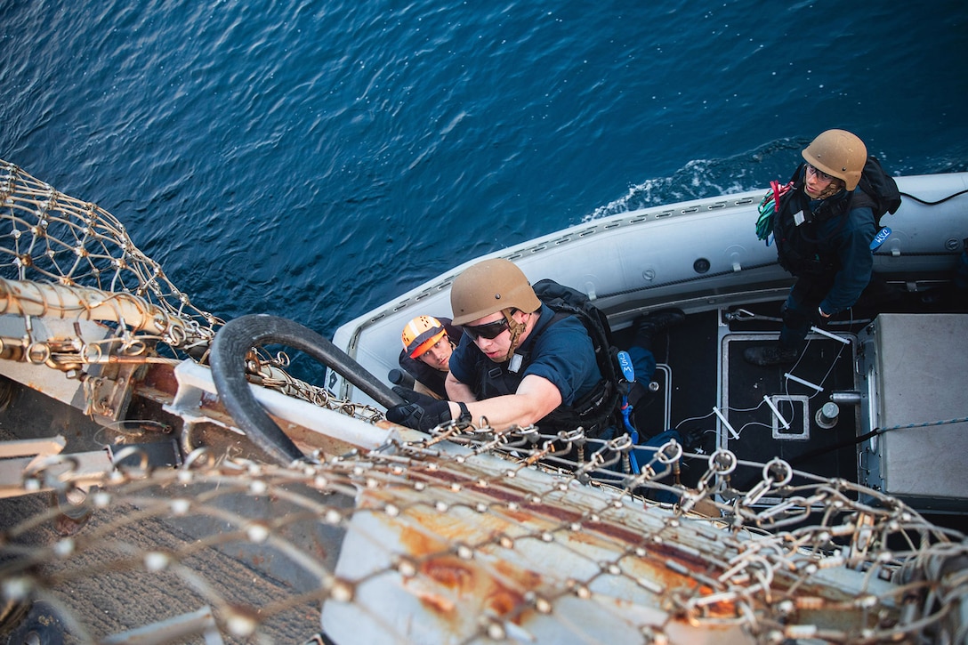 A sailor uses a hook to pull up onto a ship from a smaller inflatable boat during training as two others watch.