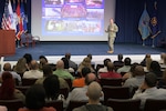 a man in uniform stands on a stage in front of a large audience with a screen with pictures behind him