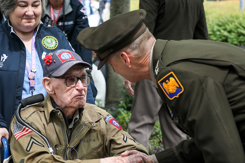 A uniformed service member leans forward to shake hands with a World War II veteran seated in a wheelchair.