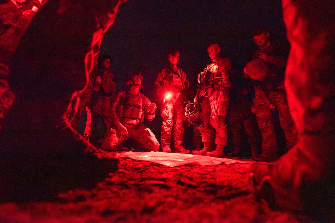 An airman kneels next to a board as cadets gather in a circle in the dark as one of them shine a flashlight.