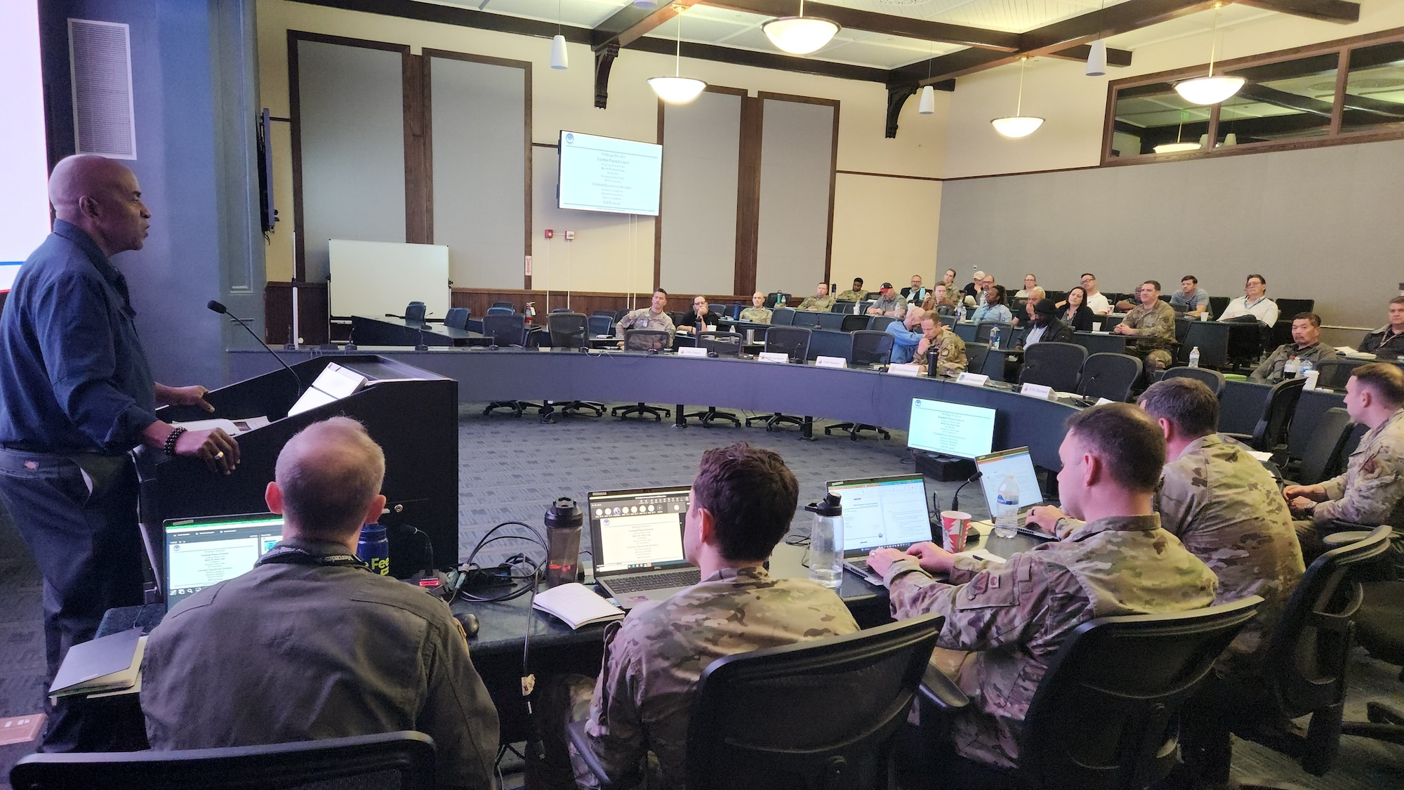 photo of large conference room with slide on wall, military members sitting at tables with computers