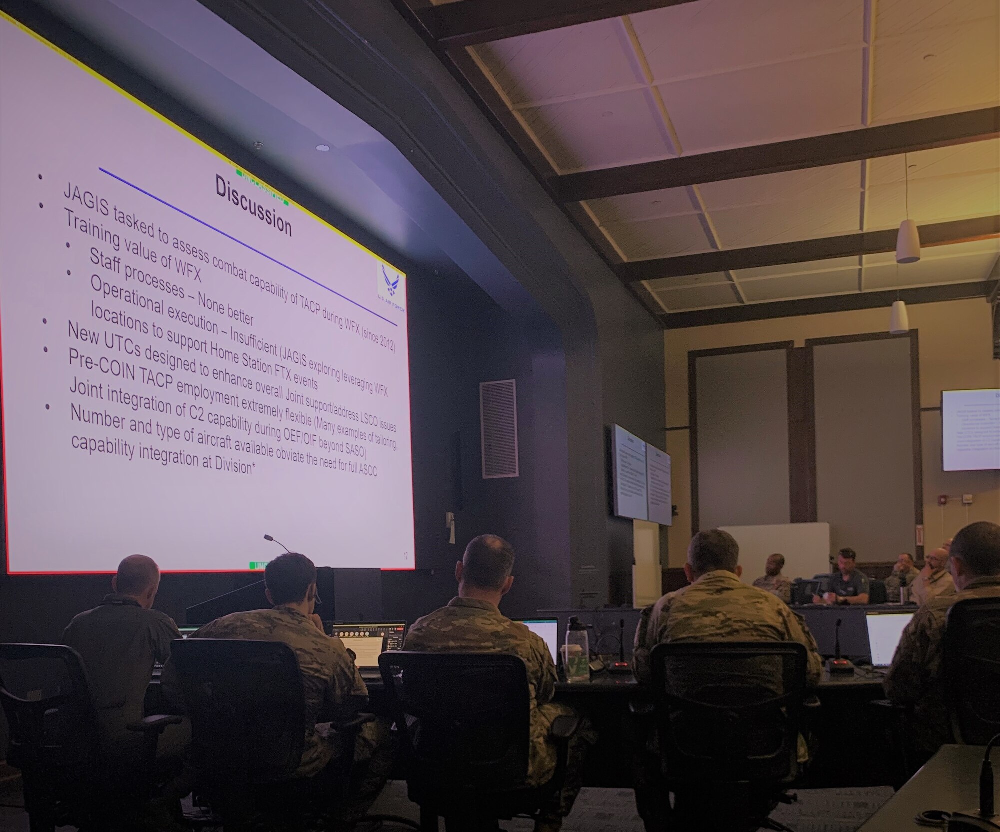 photo of large conference room with slide on wall, military members sitting at tables with computers