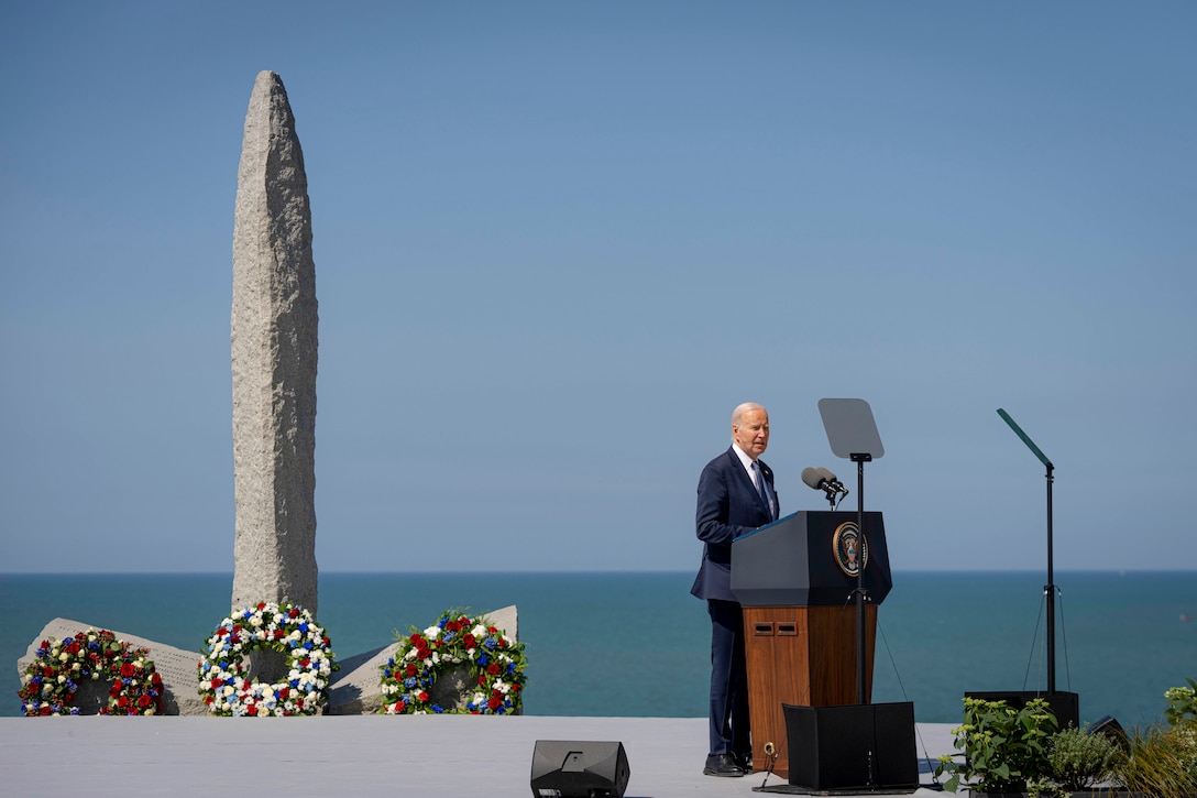 President Joe Biden stands and speaks at a lectern outdoors in front of a large granite pylon and sea in the background.