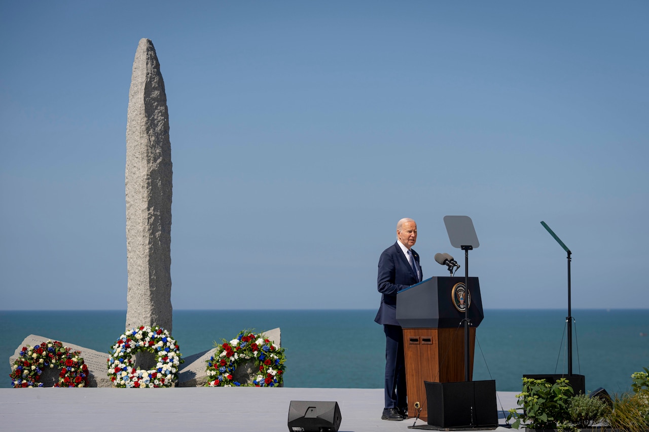 President Joe Biden stands and speaks at a lectern outdoors in front of a large granite pylon and sea in the background.