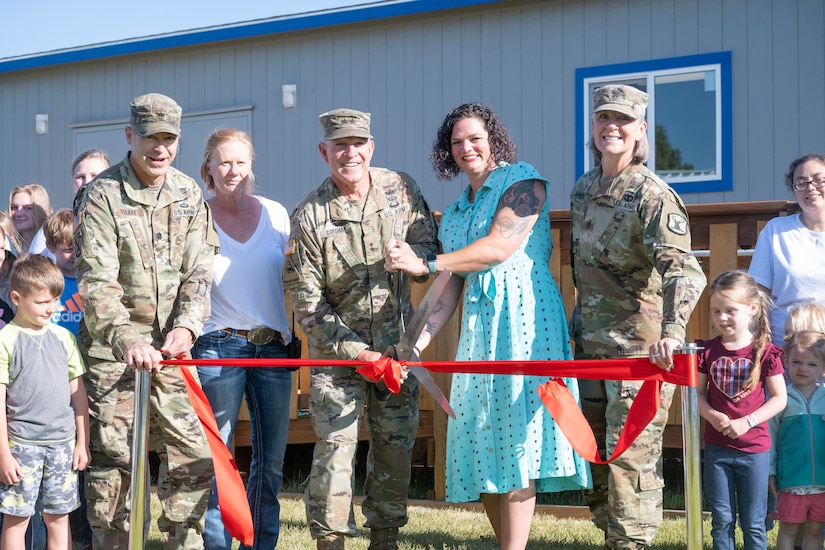 Several service members and civilians including children hold a ceremonial ribbon in front of a building while one makes a cut.