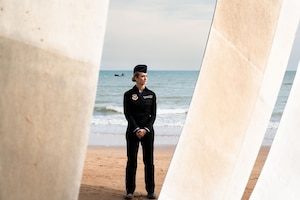 woman in "Miss America" flight suit stands in front of D-Day memorial on Omaha Beach