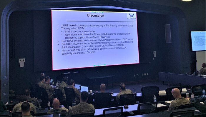 photo of large conference room with slide on wall, military members sitting at tables with computers