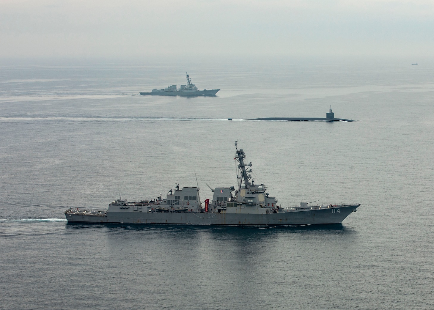 A submarine moves through the water. It is flanked by two large naval vessels.