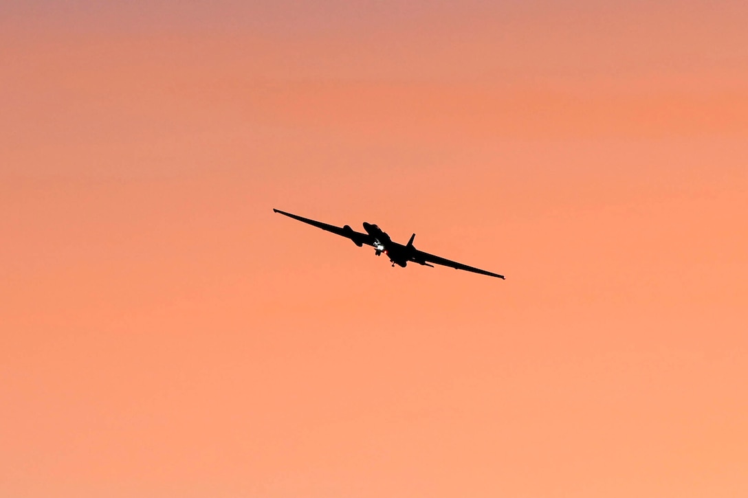 A military aircraft flies across a light orange sky.