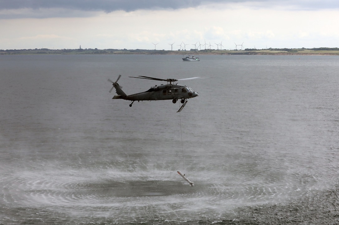 A helicopter hovers over water while lowering a cylindrical object.