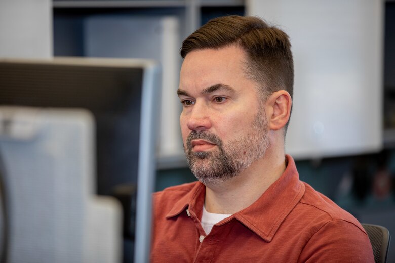 A man in a red shirt sits behind a computer.