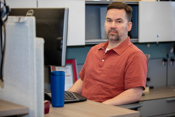 A man in a red shirt sits behind a computer with a blue coffee mug in front of him.