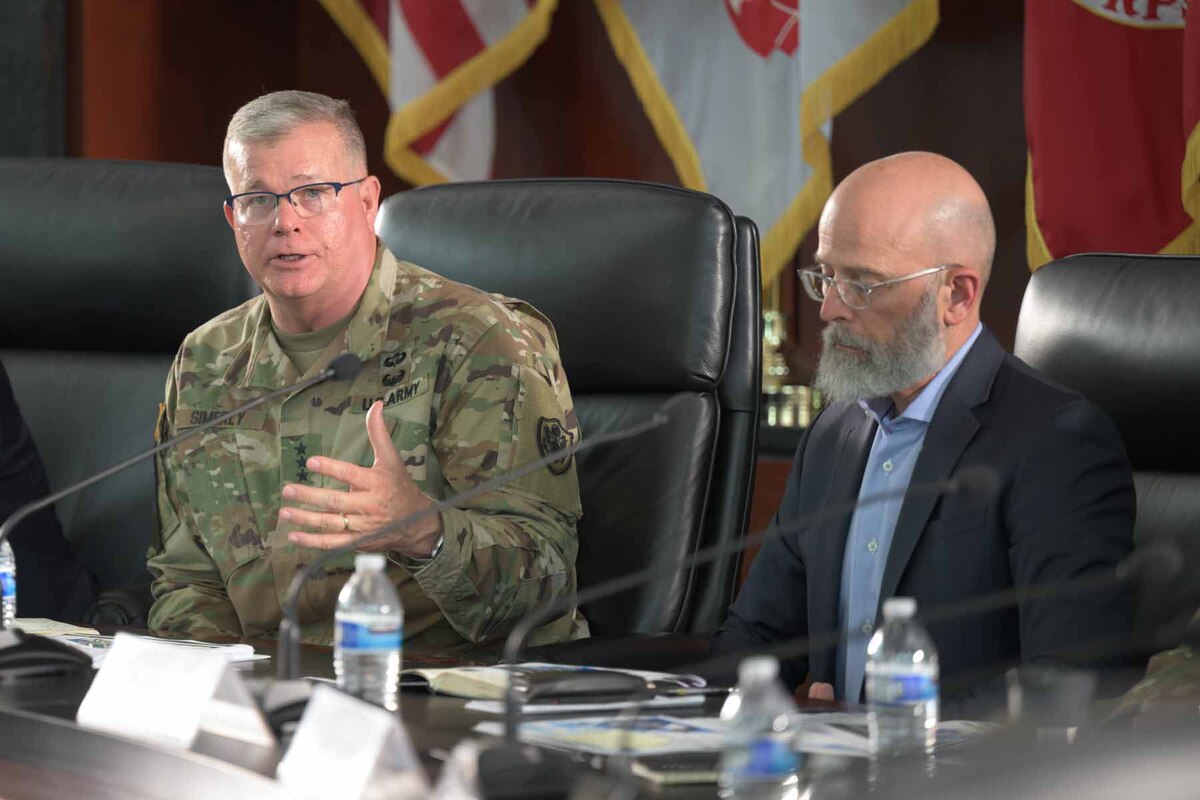 A male service member and a man dressed in civilian clothing sit at a table in front of microphones.