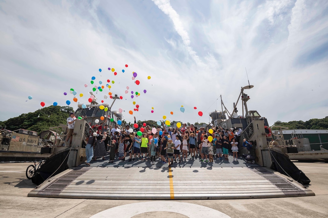 A large group of people stand near the open ramp of a docked ship and release balloons into the air.