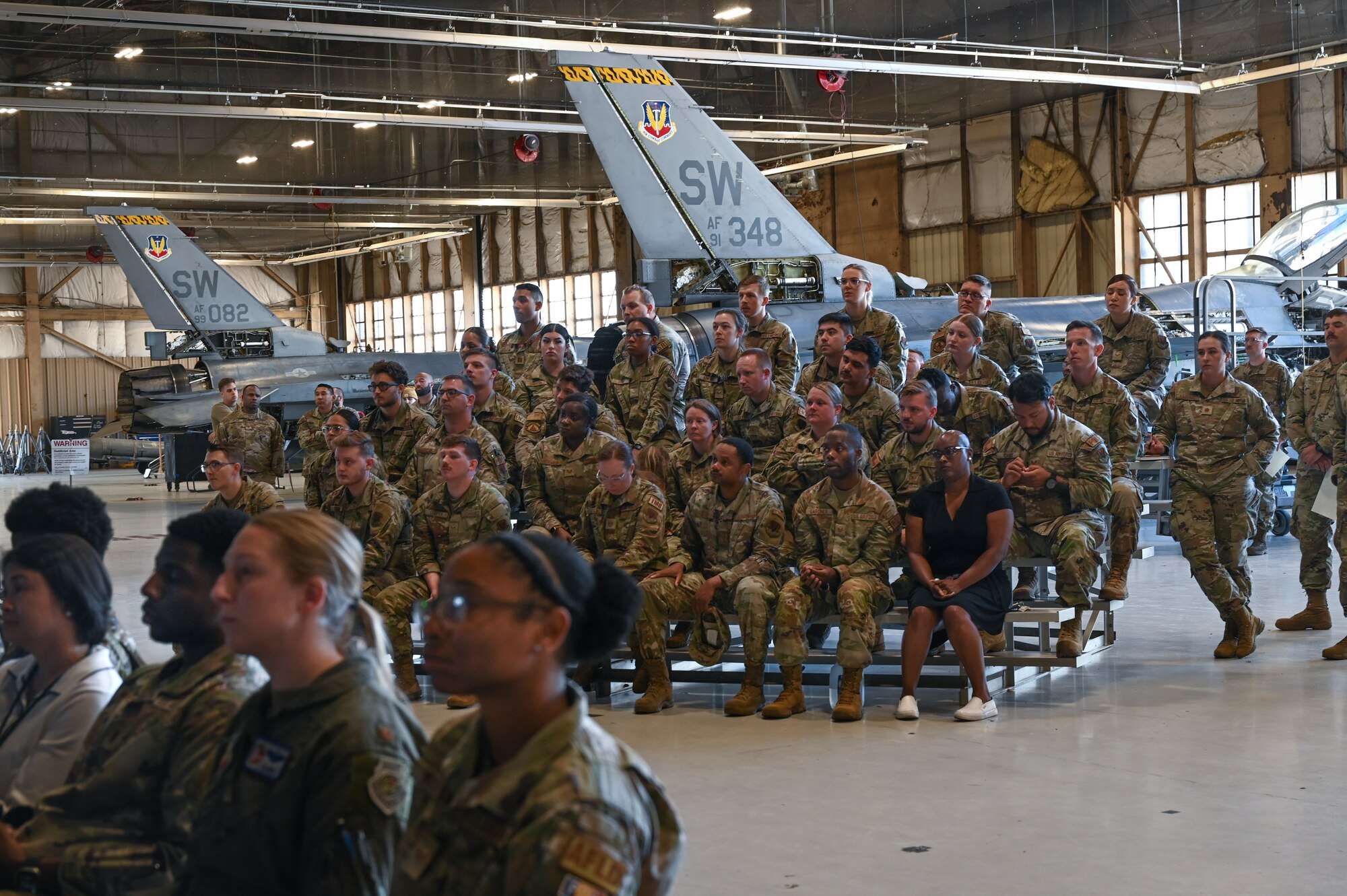 Group of military members sitting on bleachers while paying attention to the speaker.