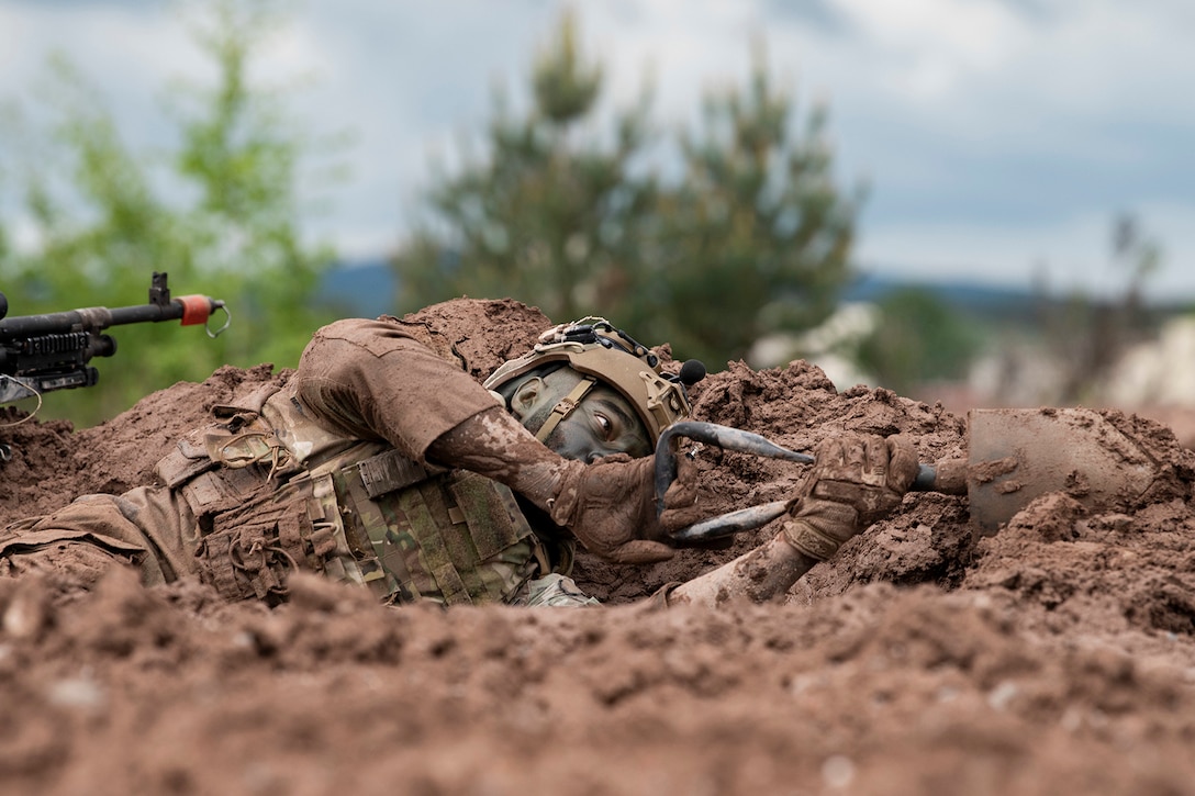 A uniformed airman lays on the ground and digs dirt with a shovel with the scope of a weapon visible on the left.