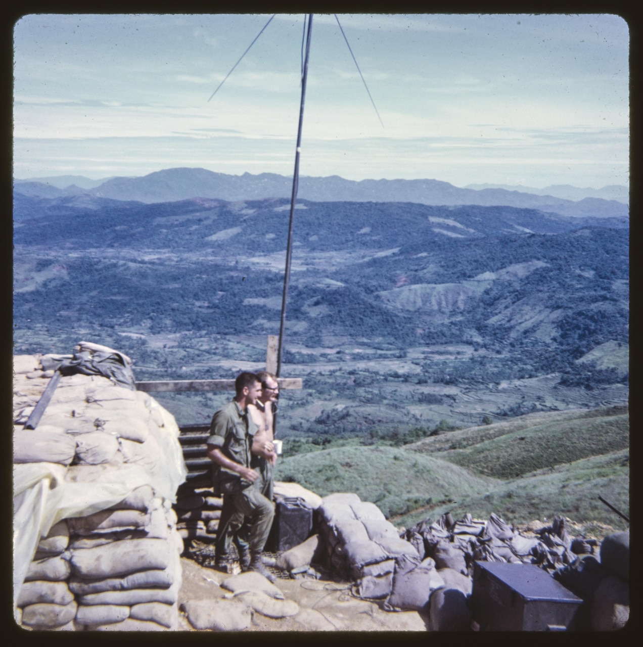 Two men stand on top of a hill that’s covered in sandbags.