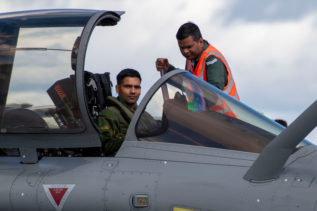 An Indian Air Force pilot looks at the camera as he sits in the cockpit of a Rafale aircraft. An Indian Air Force maintainer wearing a bright orange vest hands the pilot his bag.