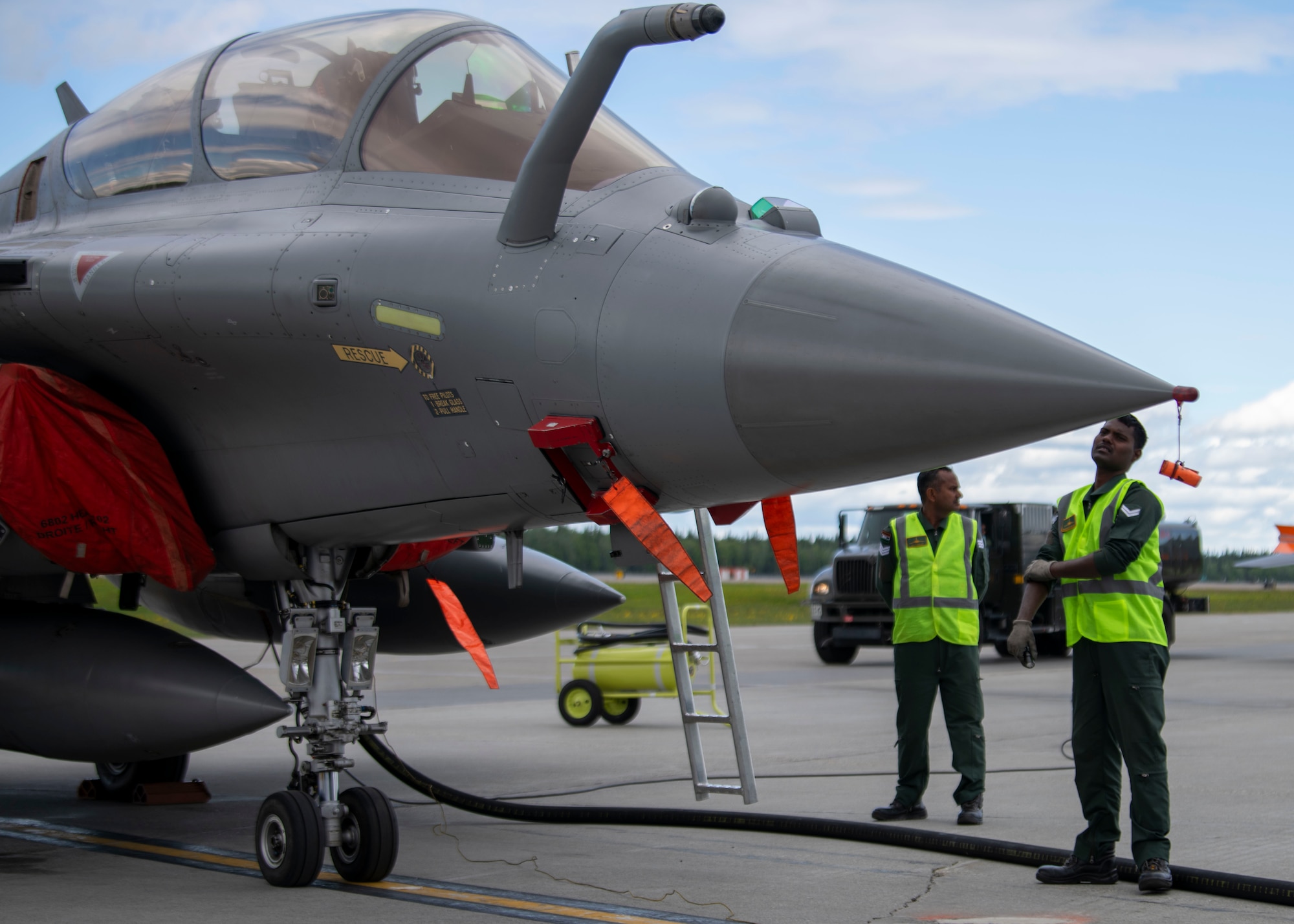 Two maintainers from the Indian Air Force stand right of a Rafale aircraft. Both maintainers are looking up at the pilot in the cockpit of the aircraft.