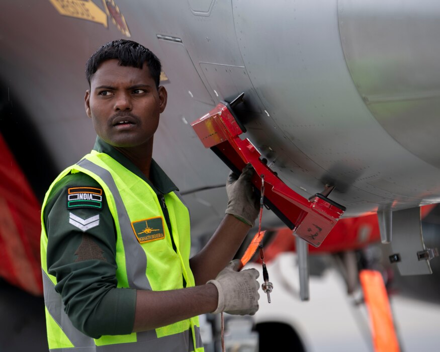 A maintainer from the Indian Air Force holds a tool against the bottom side of a Rafale aircraft.
