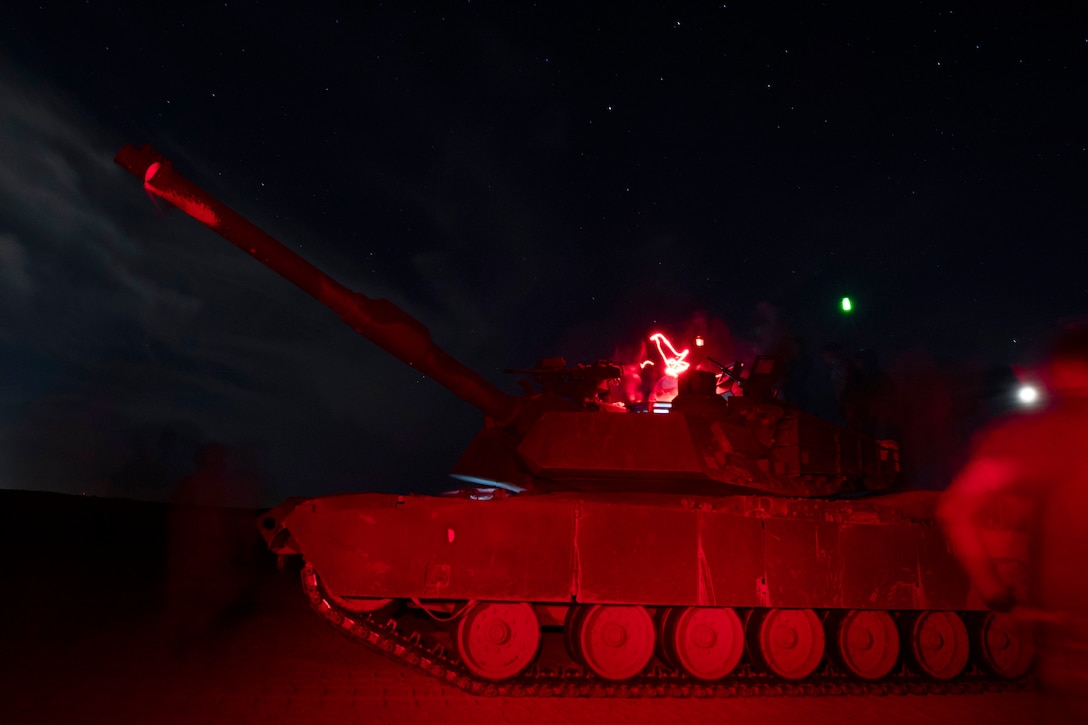 Soldiers operate a tank in the dark illuminated by a red streak as a fellow blurred soldier watches from the right.