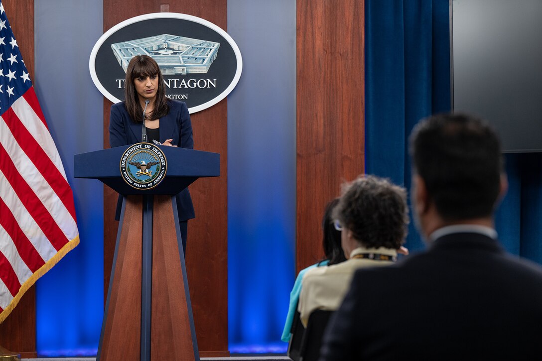 A person in business attire stands at a podium as others watch from seats.