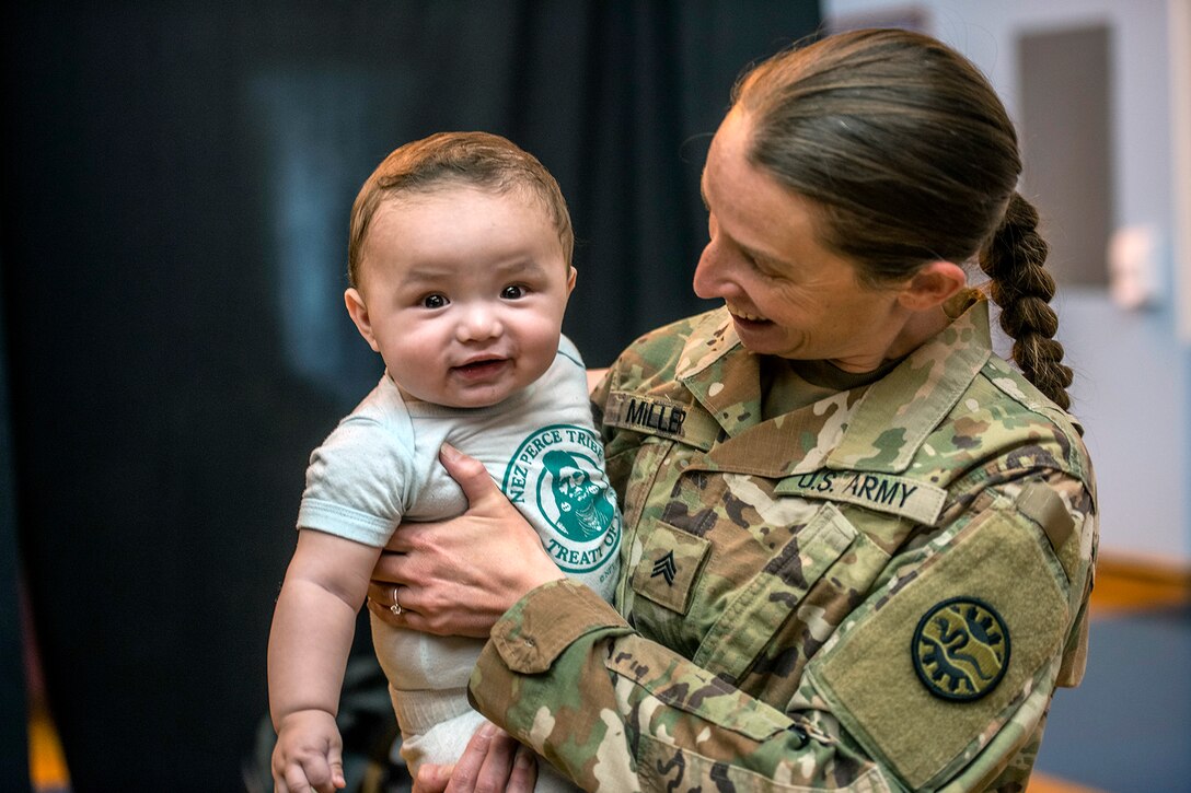 A soldier smiles at a baby they are holding while the baby smiles at the camera.