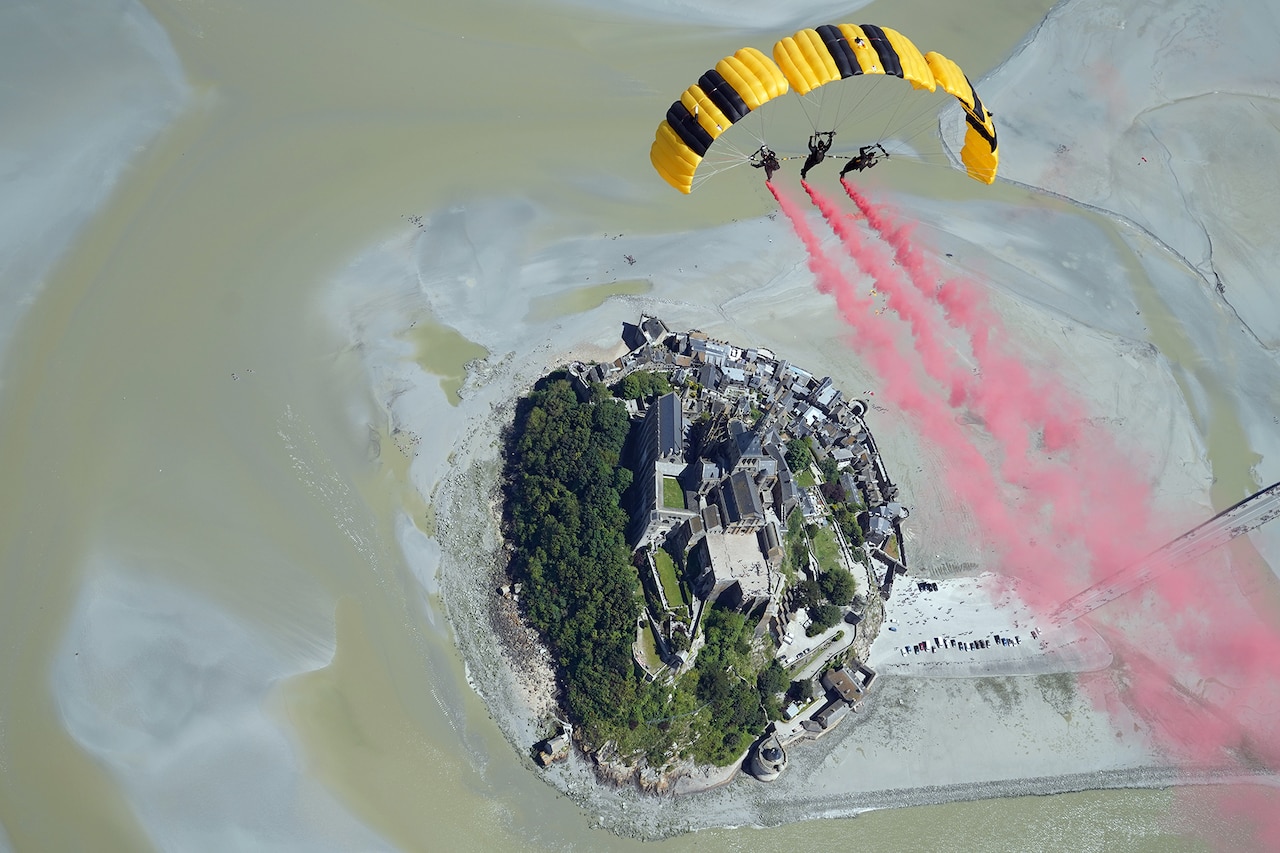 Three soldiers form a half-circle with their open parachute canopies and leave trails of pink smoke as they soar above an island.