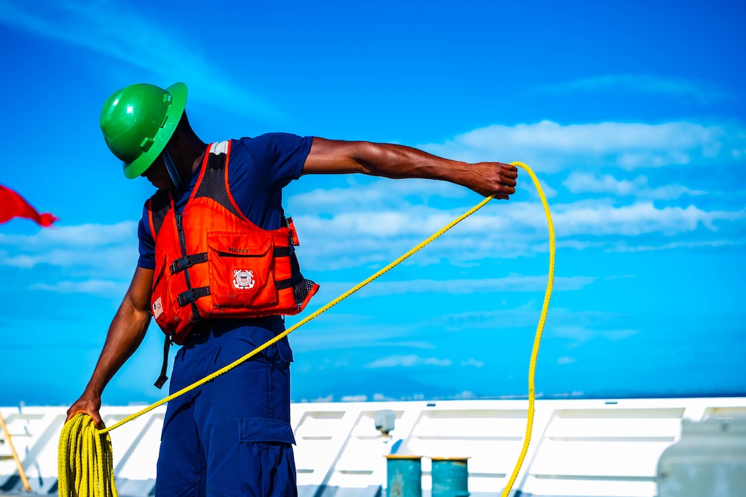 A Coast Guardsman wearing a hard hat and life vest coils up a yellow rope.