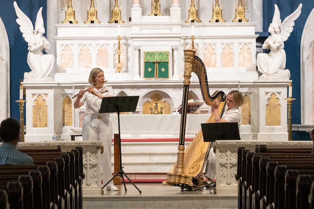 Two sailors in formal uniforms play a flute and a harp on a stage.