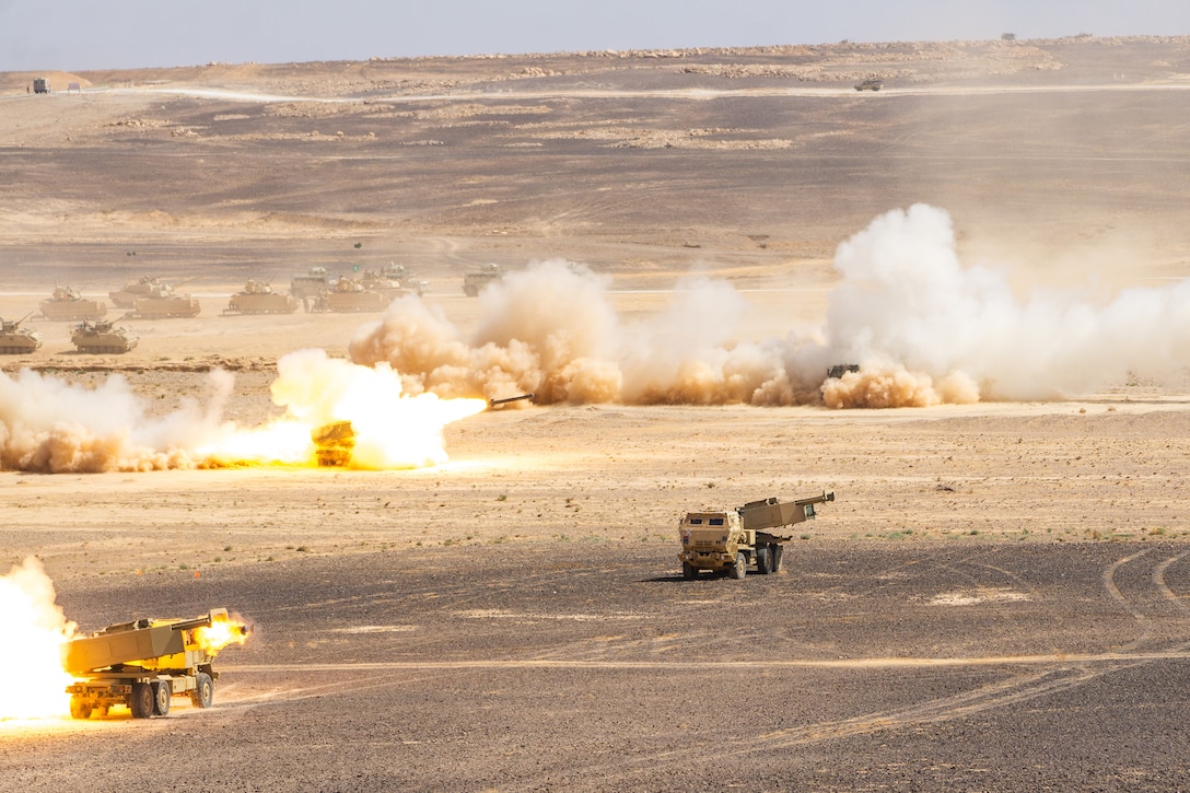 Military tanks fire rocket systems in the desert, creating dust and flames.