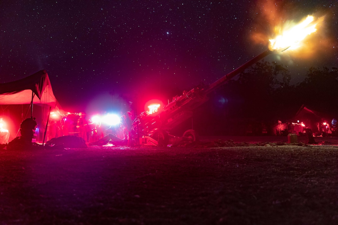 U.S. Marines and Australian soldiers standing near a tent illuminated by red and blue streaks fire a weapon under a starry night sky.