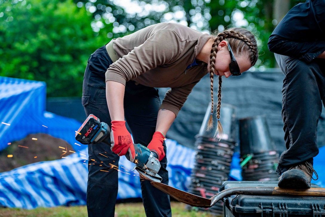 Sparks fly as a soldier bends over to sharpen a shovel while a person puts their foot on the handle on a digging site.