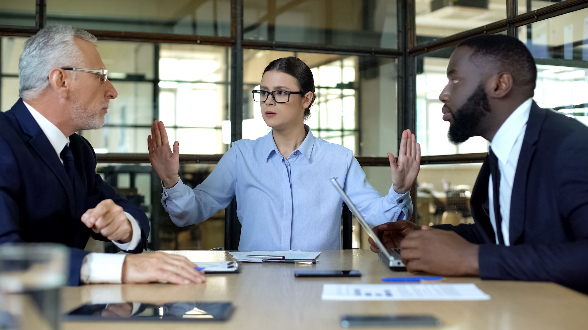 A manager holding hands up to interrupt two arguing employees sitting at a desk.