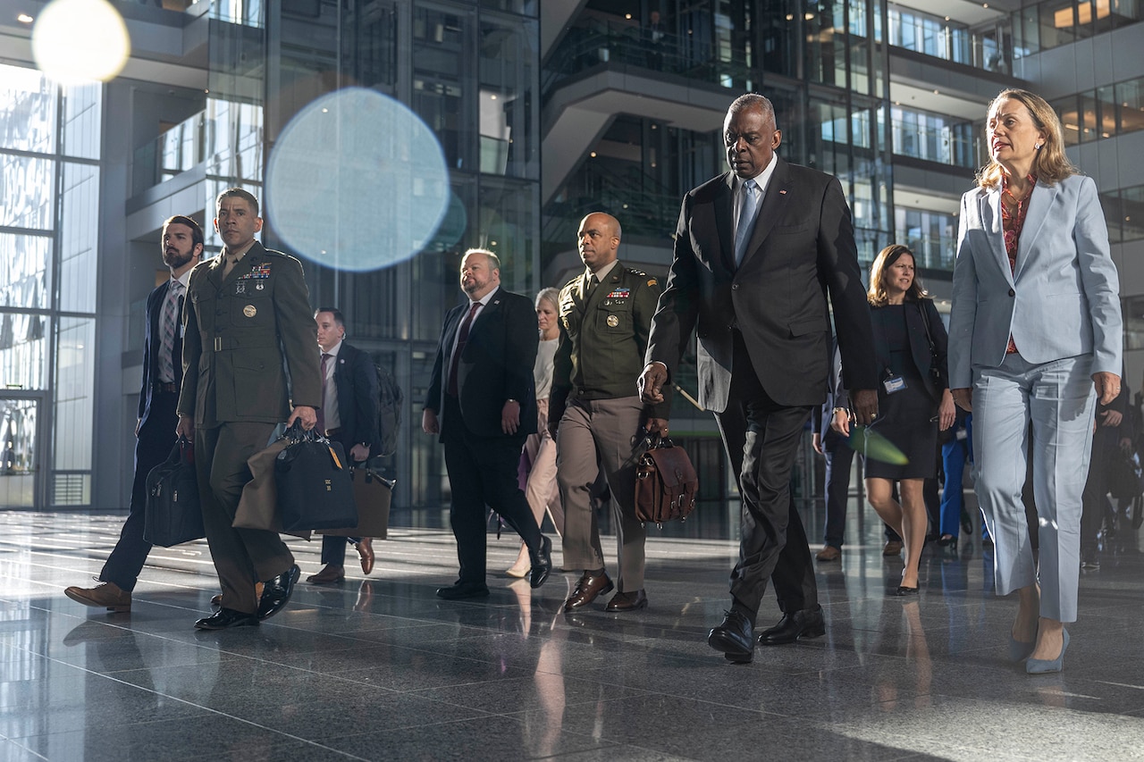 Men and women in business attire and military uniforms walk through a lobby of a building.
