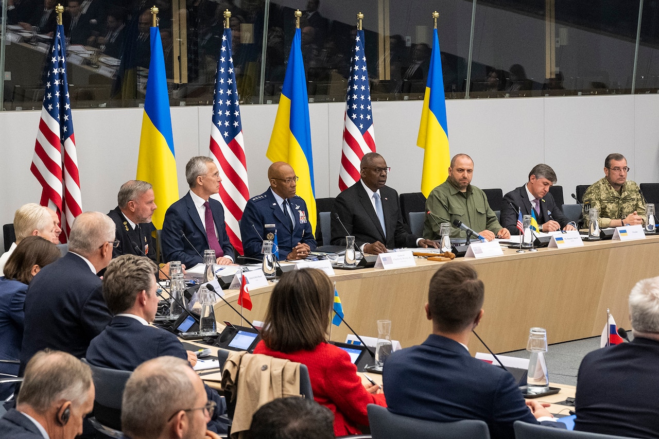 A man in business attire sitting at a conference table speaks to a group of men and women in business attire and military uniforms at the same table.  Behind the speaker are alternating U.S. and Ukraine flags.