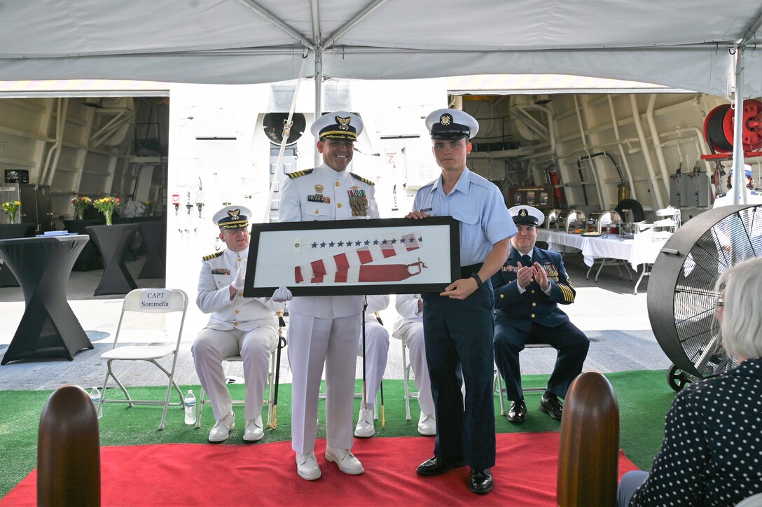 Fireman Apprentice Dylan Shira-Romero, a crew member assigned to U.S. Coast Guard Cutter Calhoun (WMSL 759), presents Capt. Timothy Sommella with the cutter’s first commissioning pennant after Sommella was relieved as commanding officer during Calhoun’s change-of-command ceremony, June 11, 2024, in North Charleston, South Carolina. The change-of-command ceremony is a time-honored military tradition that marks a transfer of total authority and responsibility from one individual to another. (U.S. Coast Guard photo by Petty Officer 3rd Class Amber Mcgruder)