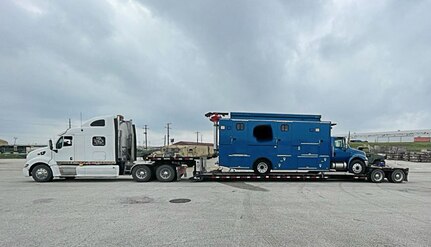 A large blue van sits on a flat bed trailer attached to a white cab semi. sitting in a parking lot. no people are in the photo.