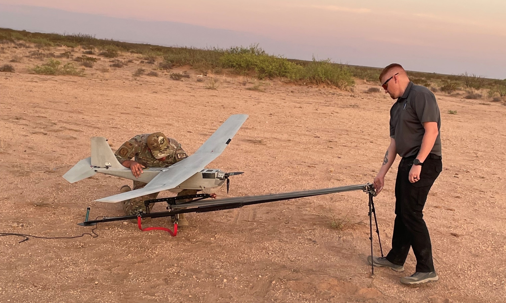 two people in the desert - one works on the sUAS while the other stands near the end of the launch platform