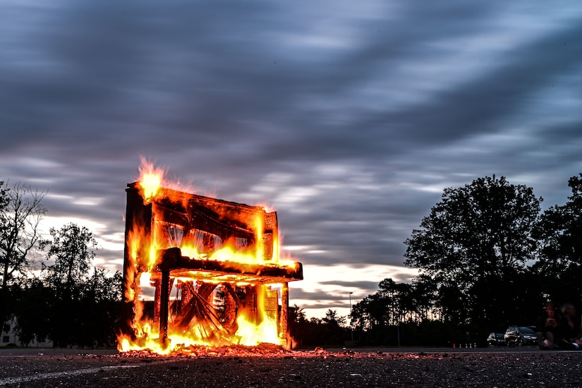 A large piano burns under a partly cloudy sky.