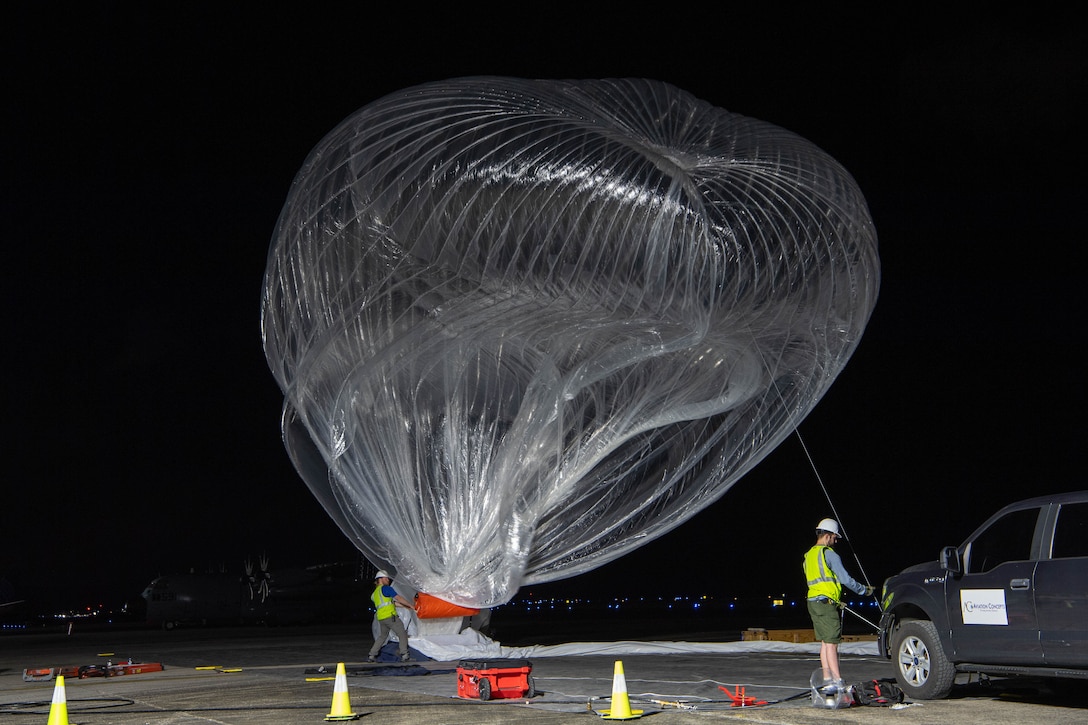 A large clear balloon floats on a tarmac at night as workers prepare it for launch.