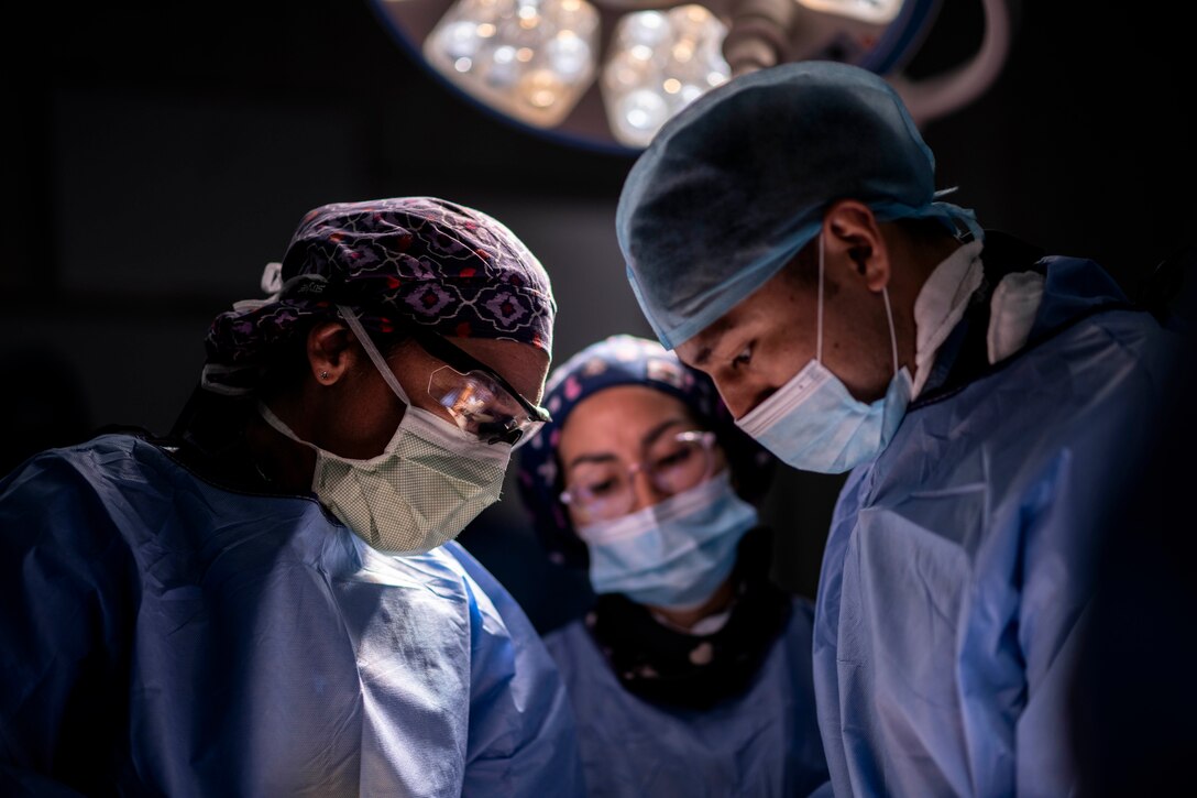 Three people wearing surgical uniforms and masks look down in a darkened room.